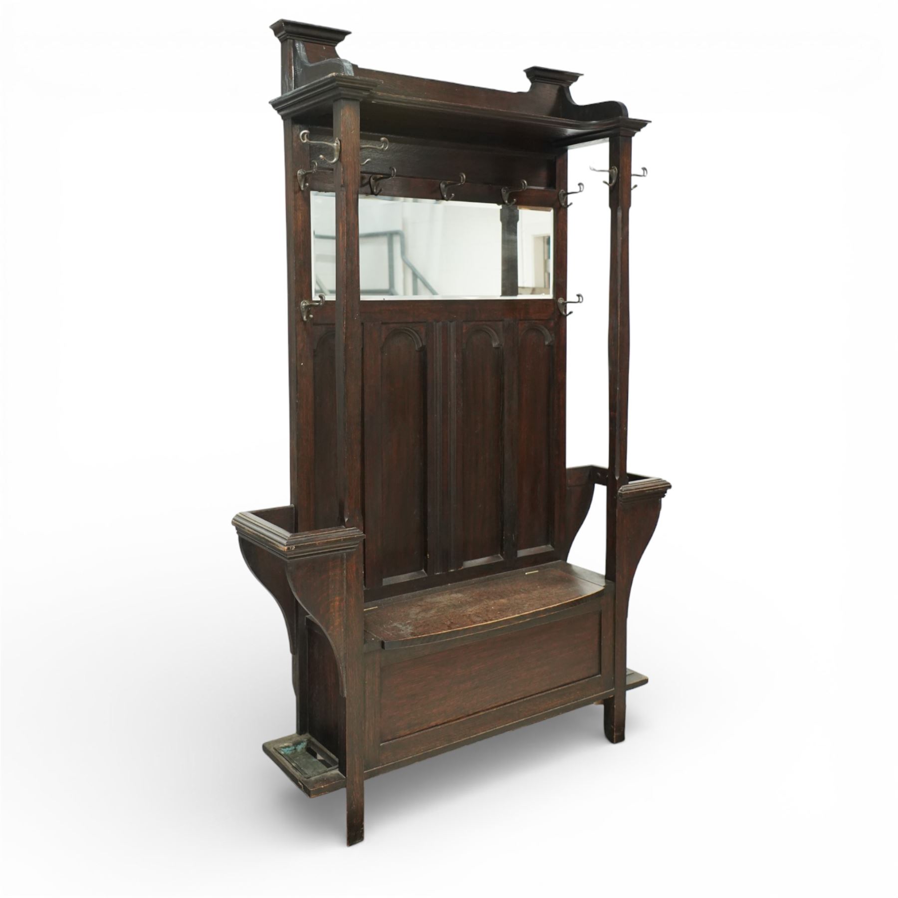 Arts and Crafts design oak settle, with projecting moulded cornice over a boarded back fitted with a rectangular bevelled mirror plate and a row of wrought iron coat hooks, the panelled back with arched fielded panels, flanked by shaped umbrella or stick stands with removable drip trays, the plank hinged seat over a panelled box base, on square section supports