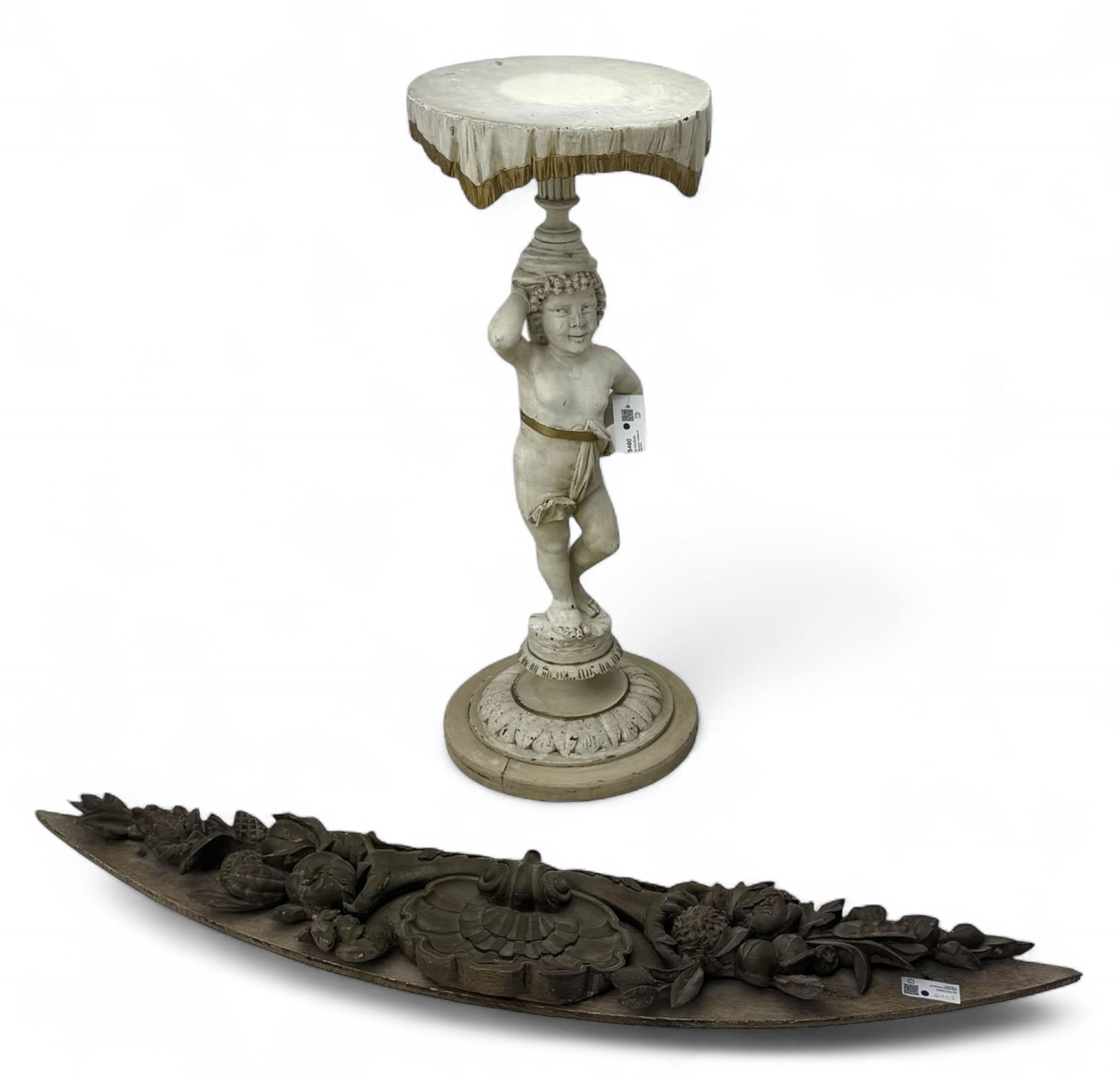 Early 20th century figural torchère, modelled as a standing putto supporting a circular top with tasselled drapery effect, raised on a circular base with foliate moulding; together with a 19th century large carved wood fragment, triangular form with high relief fruit, foliage and shell motif (H23cm, W129cm)
