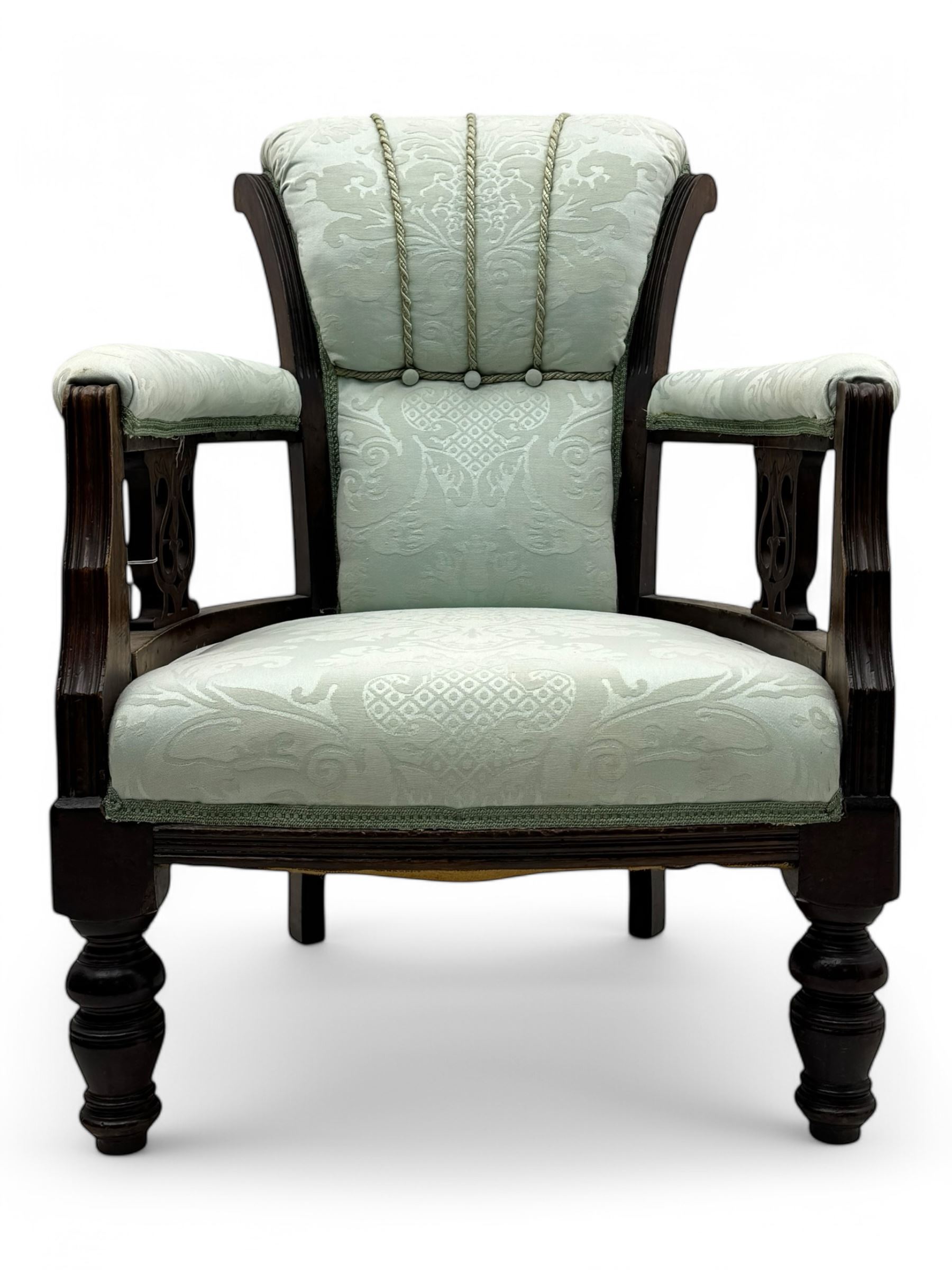 Late Victorian carved mahogany open armchair, the shaped back with fluted upholstered panel and padded arms above pierced scroll carved side supports, over a broad upholstered seat, on turned front supports