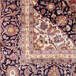 Large Central Persian Kashan hand-knotted carpet, madder red field with central cusped medallion on beige ground enclosed by indigo border with an intricate all-over arrangement of scrolling vines and floral motifs within conforming spandrels and a indigo ground main border ornamented with floral designs, enclosed by guard stripes and cotton fringe ends 