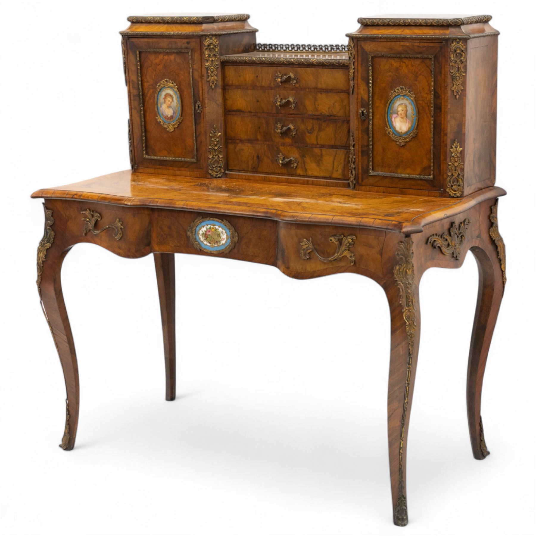 Late 19th-century Louis XV revival gilt bronze mounted and walnut Bonheur-de-Jour, rectangular-shaped serpentine form, with gilt bronze beading, frieze with two blind drawers and gilt bronze handles, centre drawer with large central serves style porcelain plaque, the elevated section with brass gallery above panelled cupboard doors also with serves style porcelain plaques and gilt bronze mounts, interior with single shelf, central section with four drawers each with gilt bronze handles, raised on slender cabriole legs fitted with gilt bronze rococo style masks and sabots