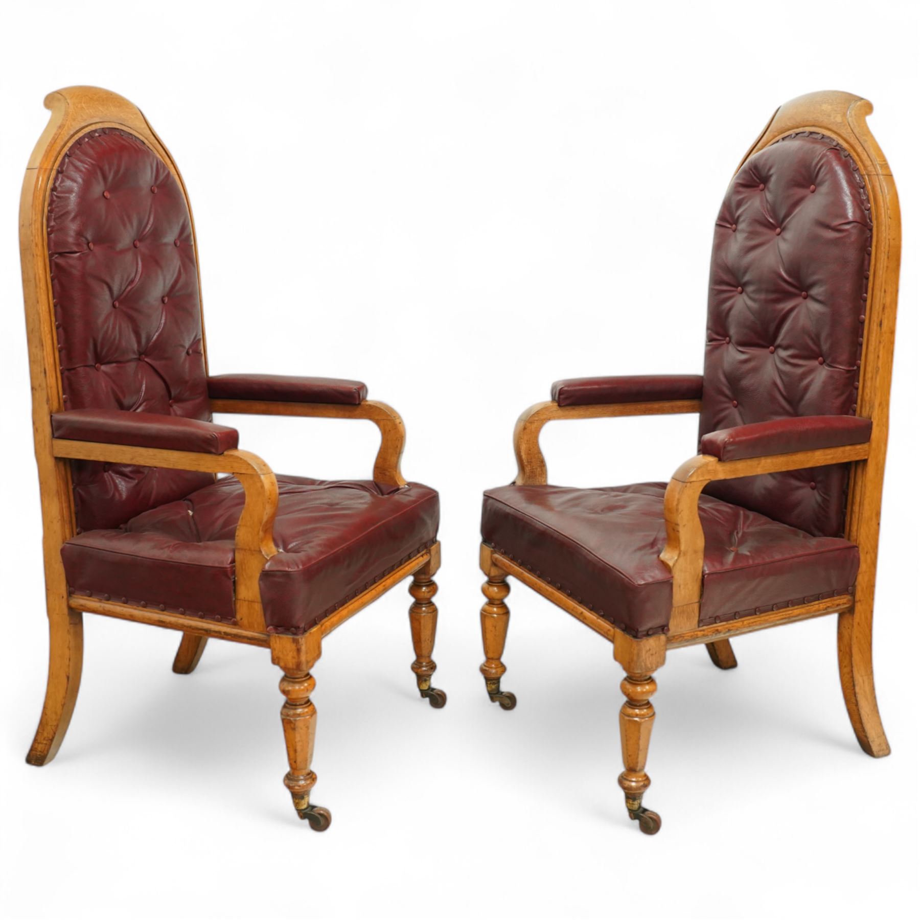 Impressive pair of mid-Victorian oak throne or hall chairs, arched backs on fluted supports with a deep buttoned upholstered panel and upholstered curved arms over fluted seat rails and tapering turned front legs with original brass and brown porcelain castors, and out-swept rear supports