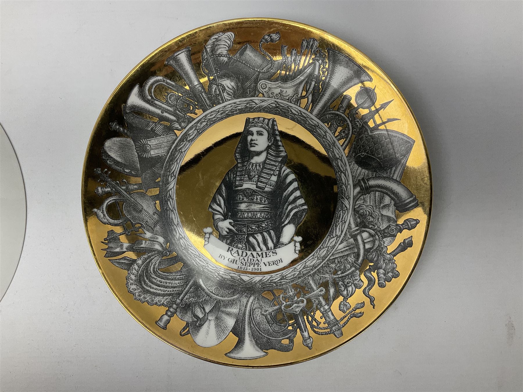Three Fornasetti plates from the Melodramma series, each depicting a operatic portraits within a border of operatic emblems on gilt ground, with printed mark beneath, D25cm 