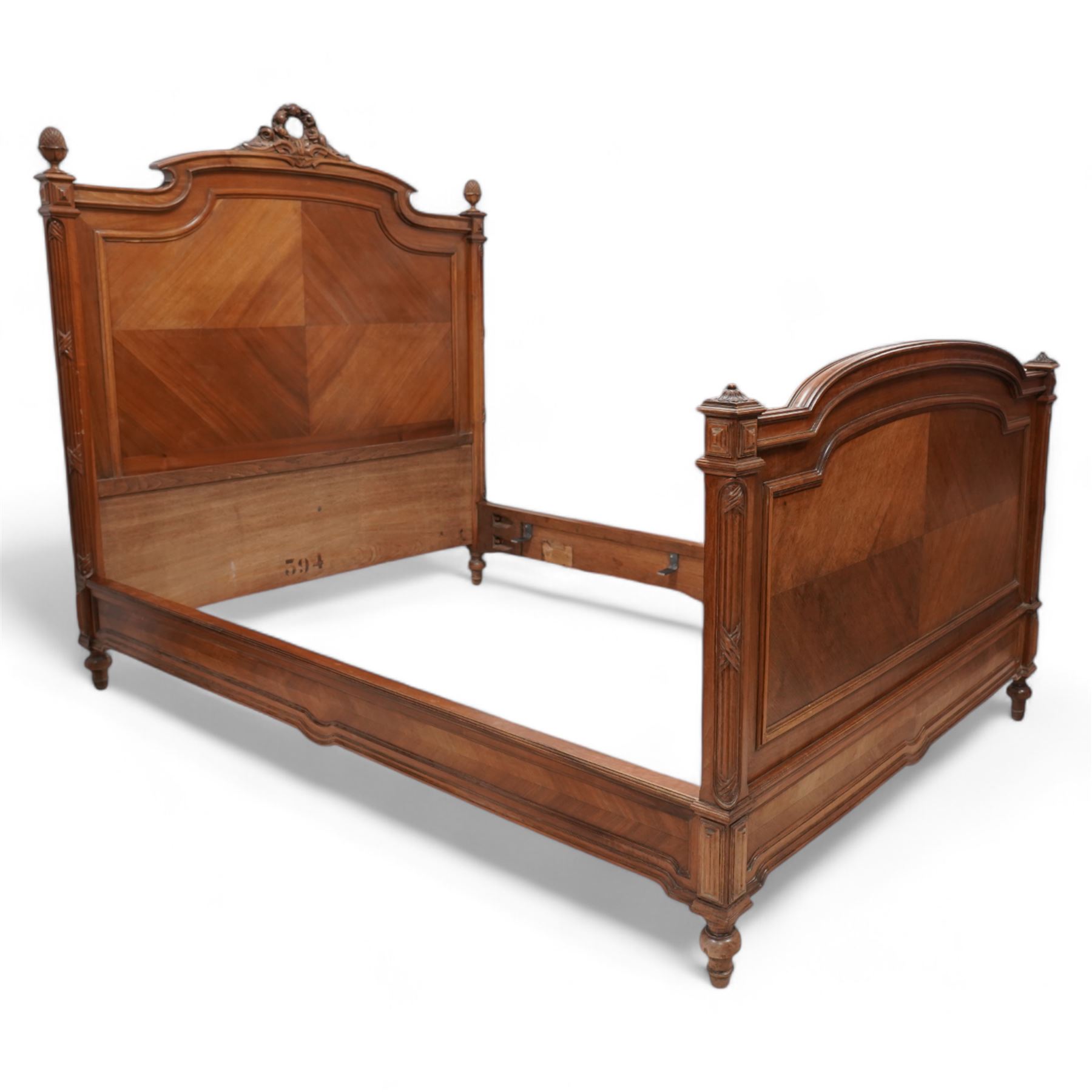 20th century French walnut 4' 6'' double bedstead, the headboard with flower head wreath pediment flanked by carved pineapple finials, reed and ribbon carved corner uprights, quarter-matched veneers, on turned feet, together with upholstered bed base