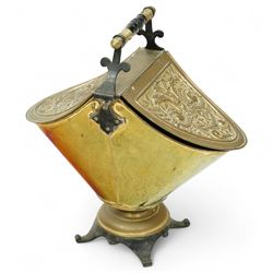 Late 19th century brass coal scuttle, of helmet form, with embossed scrolling foliate panels, cast iron supports with turned ebonised handle, raised on a cast iron tripod base