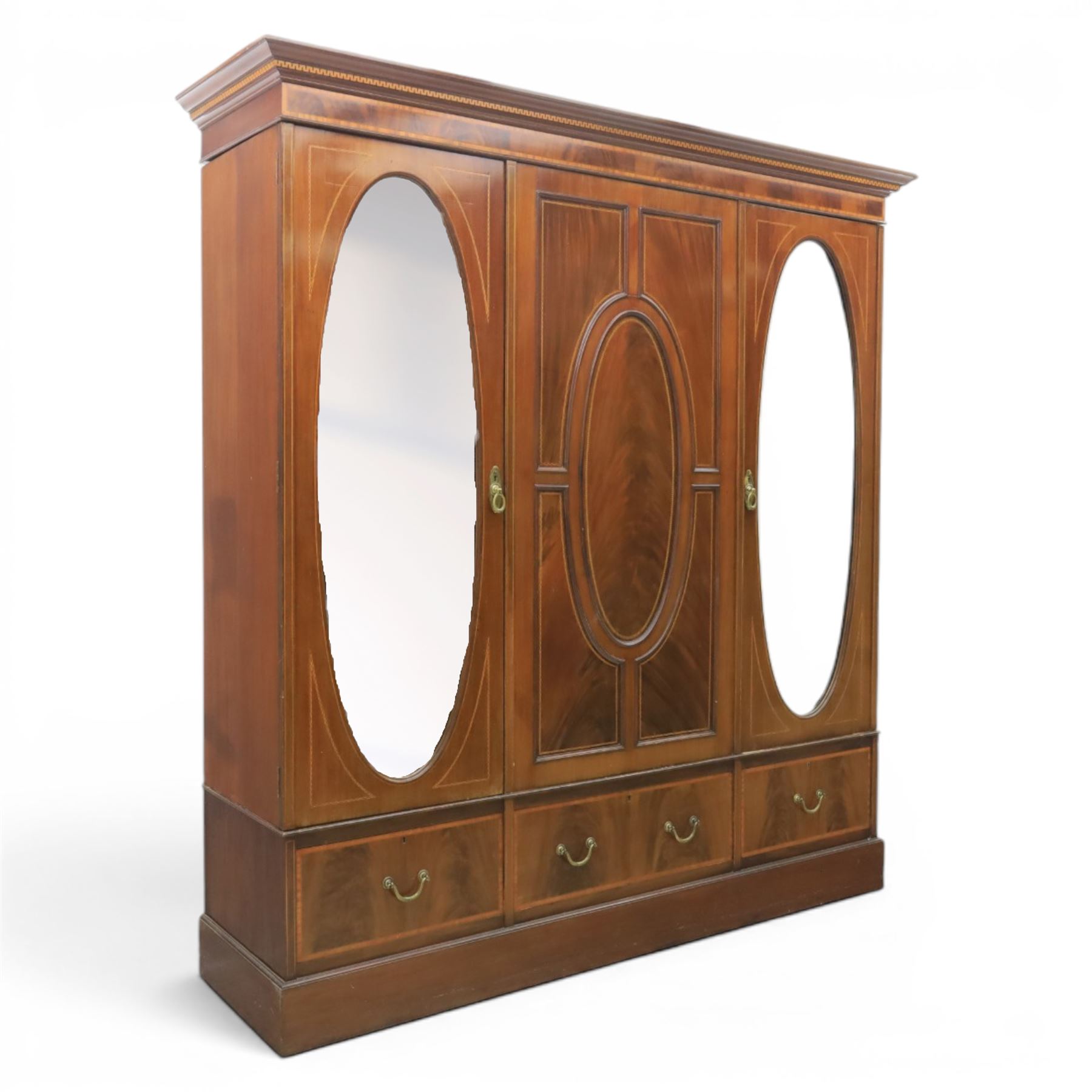 Edwardian inlaid mahogany triple wardrobe, featuring two oval mirror doors with bevelled plates, enclosing central figured veneered panel with mouled detail, above three base drawers