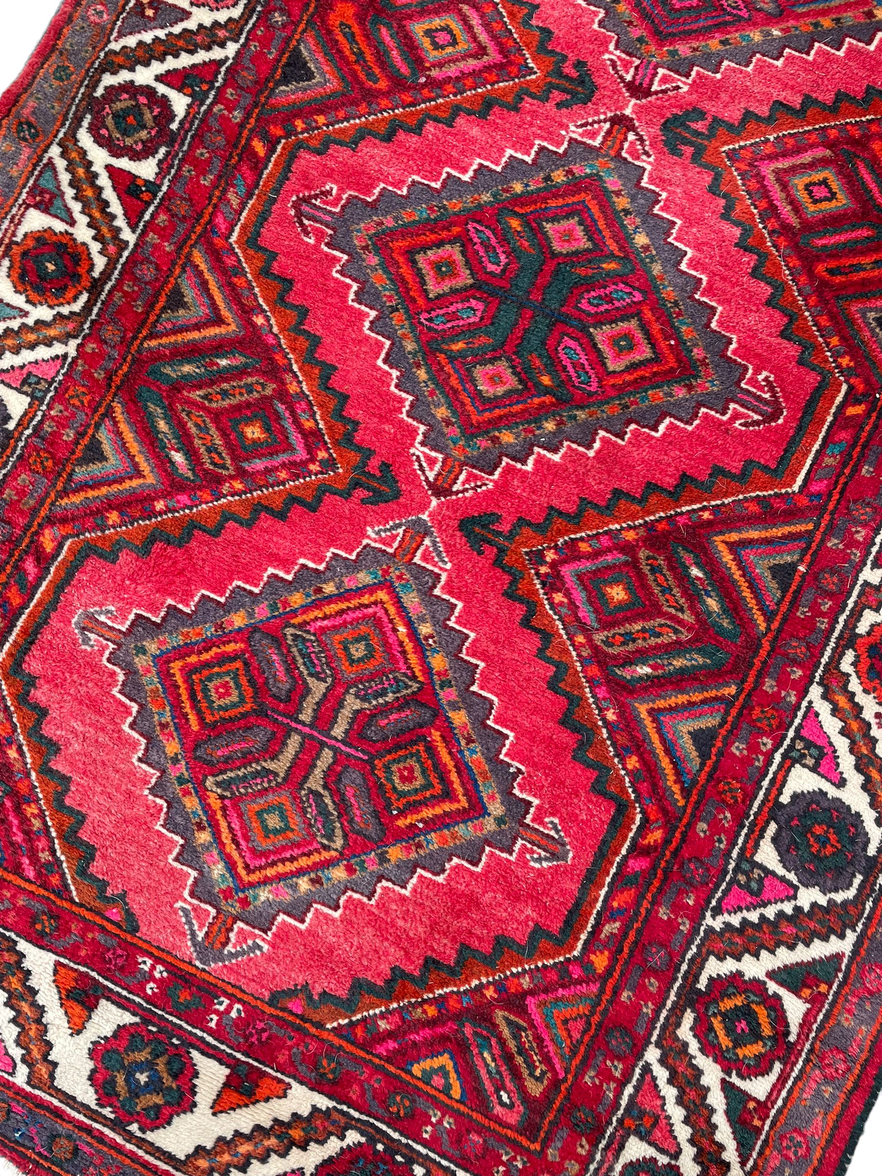 Persian Hamadan red ground rug, decorated with six stepped lozenge medallions, the guarded border with repeating stylised flower head decoration, with inscription to corner