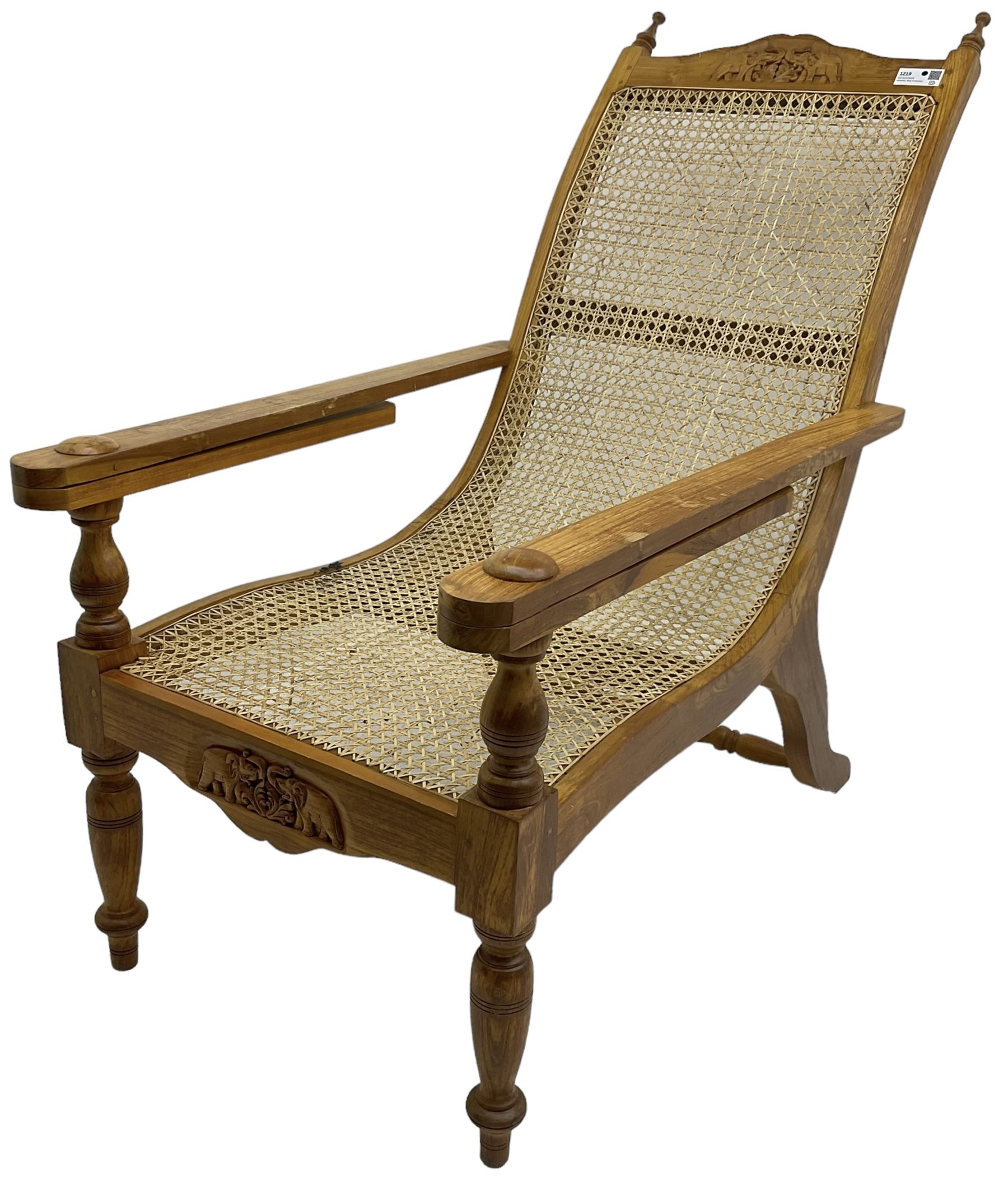 Late 20th century teak framed plantation chair, cresting rail and apron carved with two elephants with upraised trunks, flanked by finials, cane back and seat, fitted with hinged extending leg rests, on ring turned supports