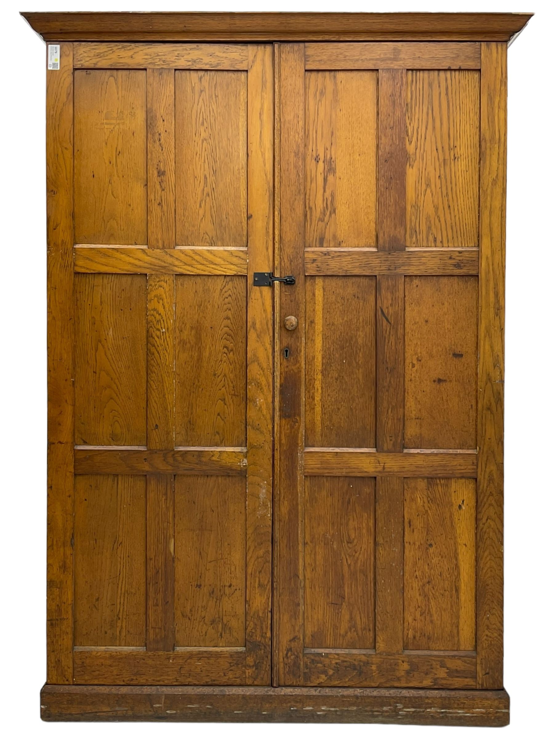 The North of England School Furnishing Company Limited Darlington - early 20th century oak school cupboard, projecting moulded cornice over two panelled doors, fitted with shelves, on chamfered plinth base 