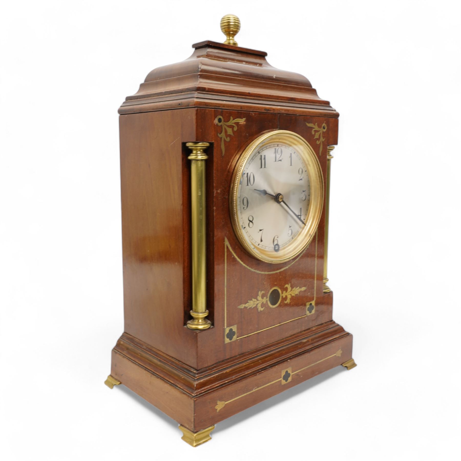 Edwardian- Early 20th century 8-day mahogany mantle clock, with a stepped pediment and brass finial, recessed brass pilasters and brass inlay to the front with a conforming plinth raised on bracket feet, silvered dial with Arabic numerals and steel spade hands within a glazed brass bezel, single train timepiece movement with pendulum and key.

