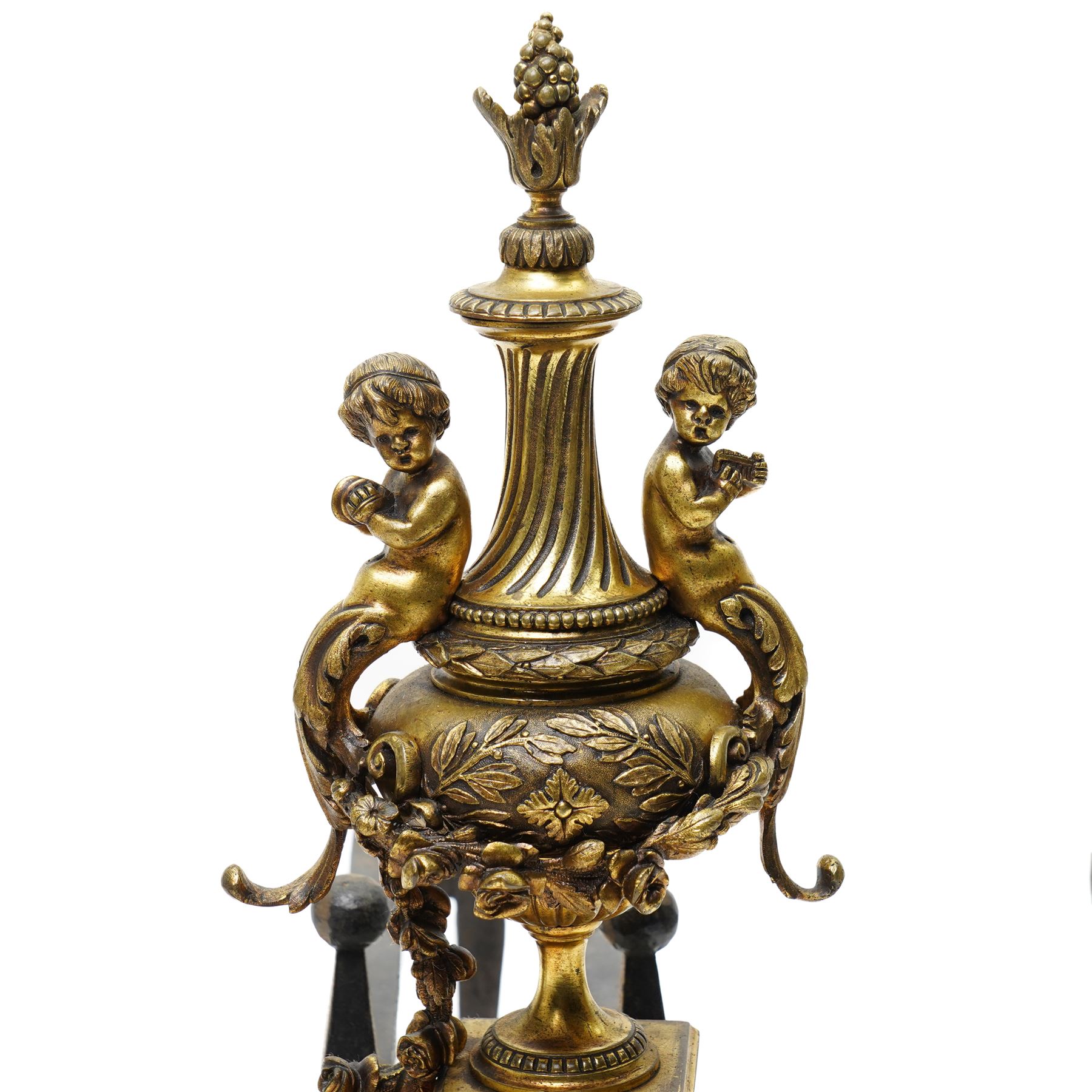 Pair of gilt metal fire dogs, formed as an urn with classical decoration, including putti and floral swags, raised on a square stepped base, the feet formed as rams heads and hoofs, H45cm x W28cm
