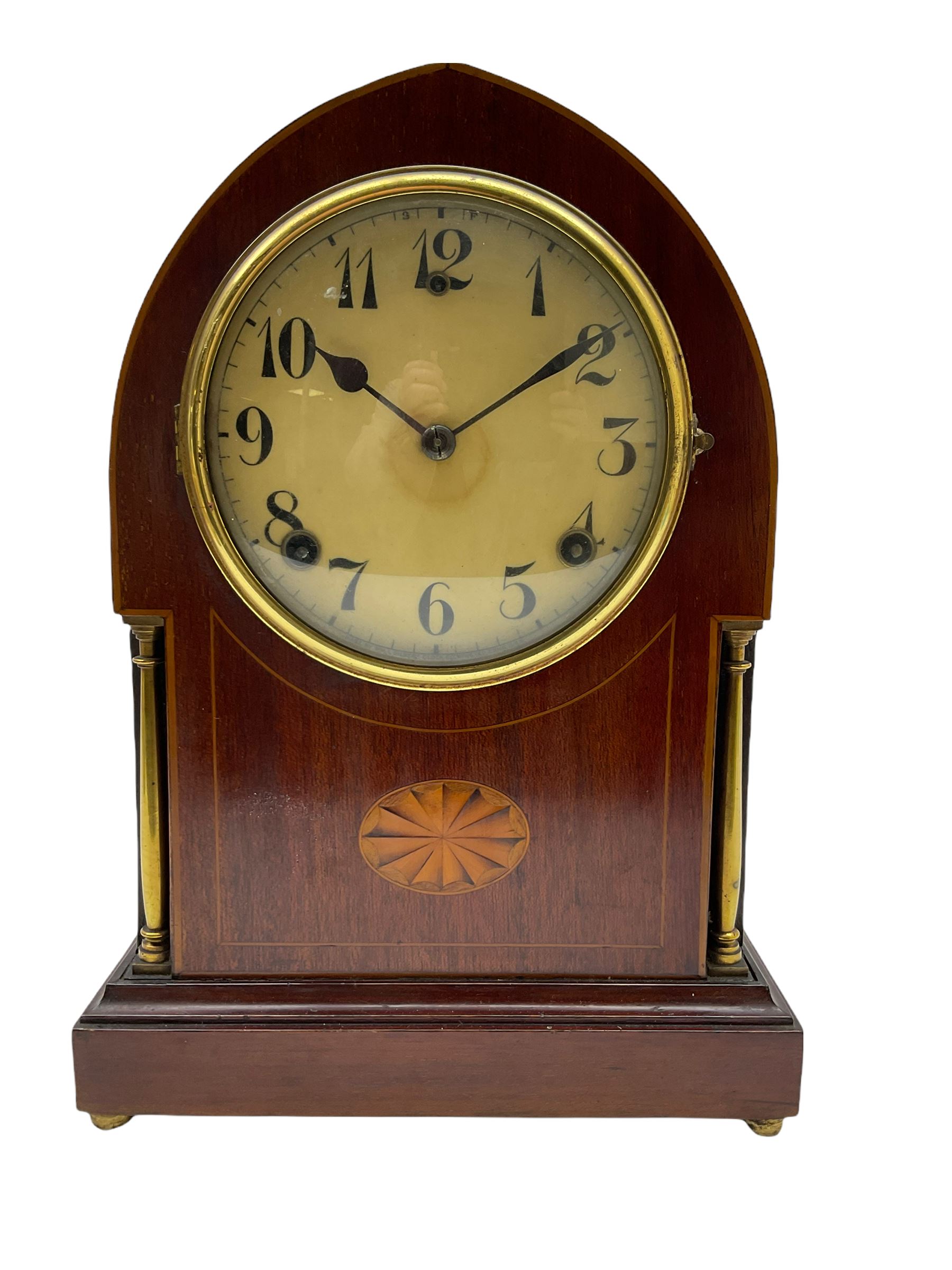 A late 19th century American  mantle clock in a “Lancet” case manufactured by the  Gilbert Clock Factory, Connecticut,  mahogany veneered case with an oval inlay, satinwood stringing to the edge and two recessed brass pillars, on a moulded plinth with four bun feet, enamel dial with upright Arabic numerals and spade hands, fast/slow regulation, with a convex glass and spun brass bezel, eight-day movement striking the hours and half hours on a coiled gong. No pendulum.