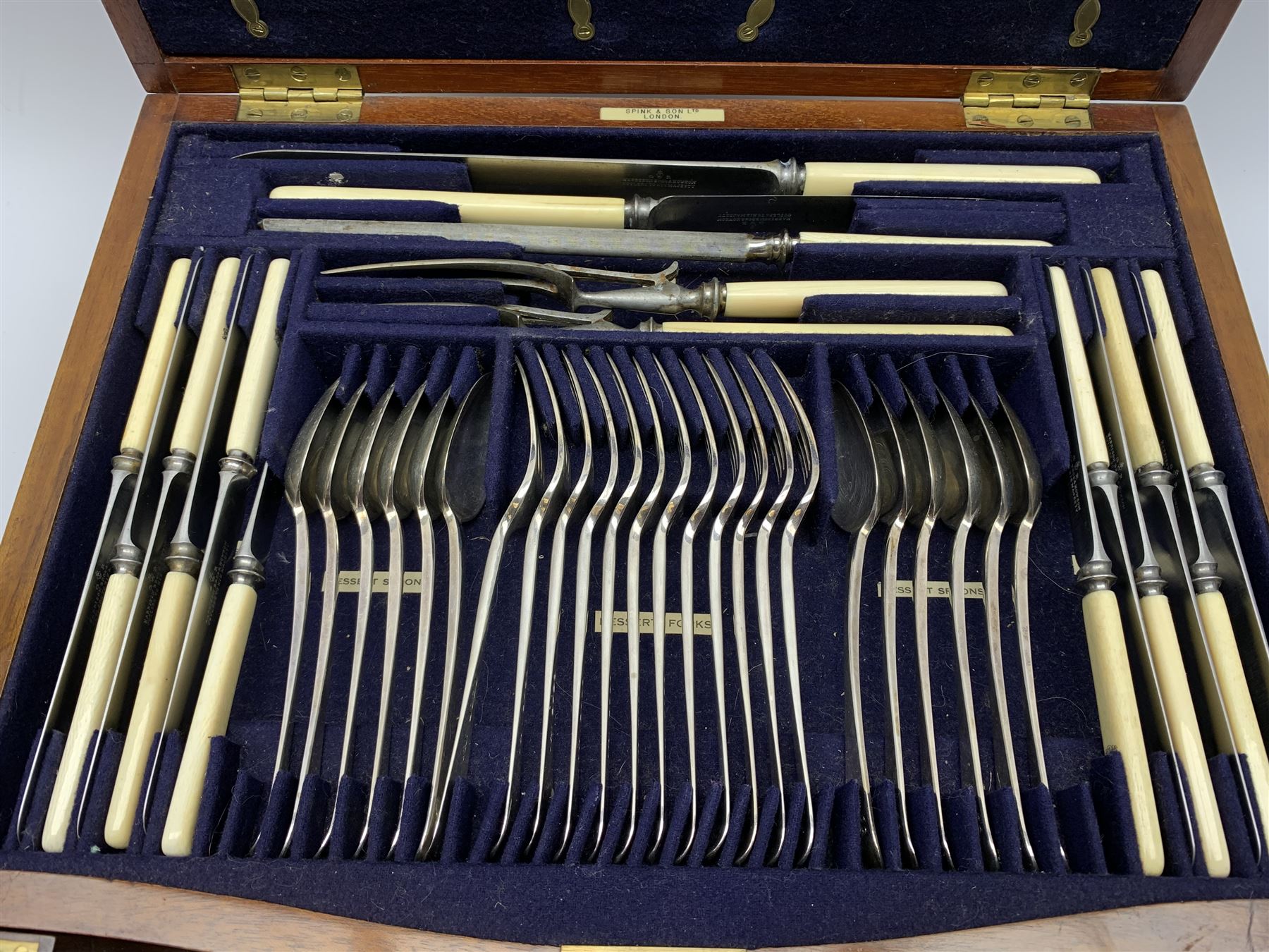 Late 19th/early 20th century composite canteen of silver flatware, contained within an early 20th century mahogany serpentine fronted case with twin drop carry handles, and hinged cover with boxwood stringing and central inlaid shell motif, above a single drawer with twin ring handles and Wellington style locking system, the cover and drawer opening to reveal plaque for Spink & Sons Ltd London, and fitted interior containing ivory handled knives, ivory handled carving set, and silver Hanoverian and Old English pattern cutlery, each with engraved monogram to terminal, hallmarked London, makers Spink & Son (John Marshall Spink), Goldsmiths & Silversmiths Co, and Chawner & Co (George William Adams), various dates ranging 1880-1923, gross weighable silver 92.59 ozt (2880 grams)