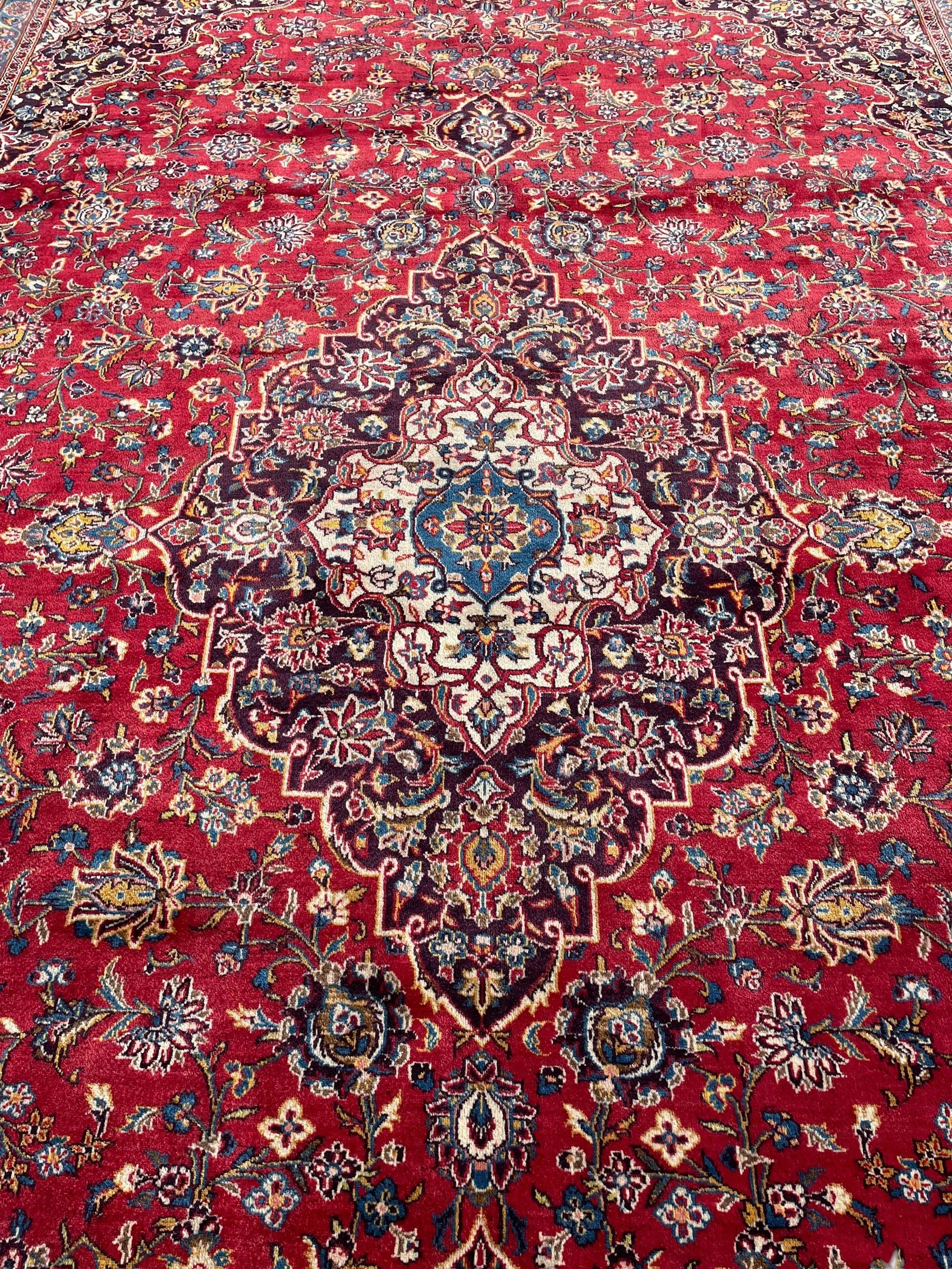 Large Persian Kashan crimson ground carpet, large floral central medallion surrounded by trailing branches decorated with leaves and stylised plant motifs, the main border with scrolling design interspersed with palmettes, within floral pattern guard stripes 