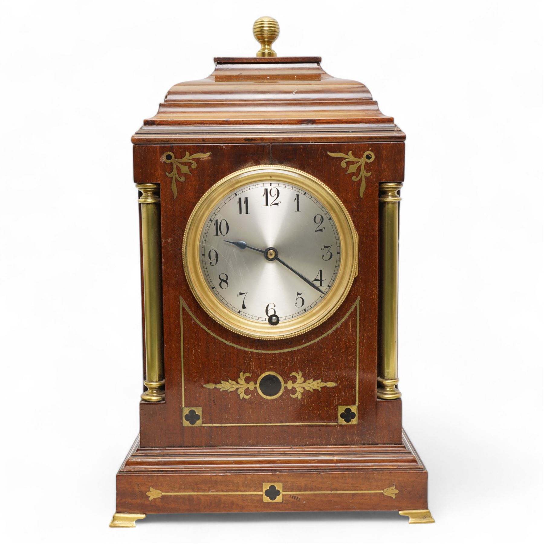 Edwardian- Early 20th century 8-day mahogany mantle clock, with a stepped pediment and brass finial, recessed brass pilasters and brass inlay to the front with a conforming plinth raised on bracket feet, silvered dial with Arabic numerals and steel spade hands within a glazed brass bezel, single train timepiece movement with pendulum and key.
