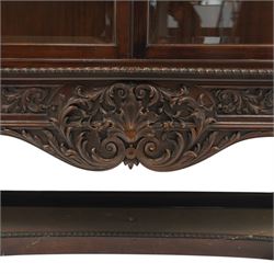 Large and impressive 19th century heavily carved mahogany display cabinet, the ornate pediment carved with a central scalloped and foliate rocaille motif with extending acanthus leaves and egg-and-dart moulding flanked by acanthi finials, over a central cartouche with carved bow-and-arrow motif and trailing scrollwork, the bookcase enclosed by two glazed doors with bevelled glass sides, the base with a gadrooned upper edge over a central rocaille ornament with scrolling acanthus leaves, the panelled frieze with further foliate motifs, raised on large carved snarling lion mask supports with floral garlands and claw feet, united by a concave rectangular platform base with gadrooning egg-and-dart moulding