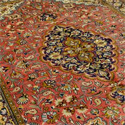 Central Persian Qom hand-knotted carpet, salmon pink ground, with a central dark navy shaped floral medallion with pendants, surrounded by dense scrolling foliate and floral motifs incorporating rosettes, palmettes and delicate vine work, enclosed by ivory spandrels and a dark navy border with repeating palmettes and meandering vines, flanked by narrow floral guard stripes, with cotton fringes