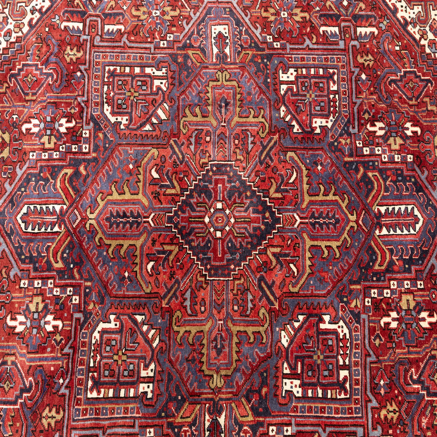 North West Persian Heriz hand-knotted carpet, madder red field with central bold angular steeped medallion with serrated outlines, flanked by stylized palmettes, hooked leaves and geometric motifs, spandrels similarly decorated within a wide geometric border and multiple guard borders in tones of madder red, indigo blue, ivory and olive green, with cotton fringe ends  