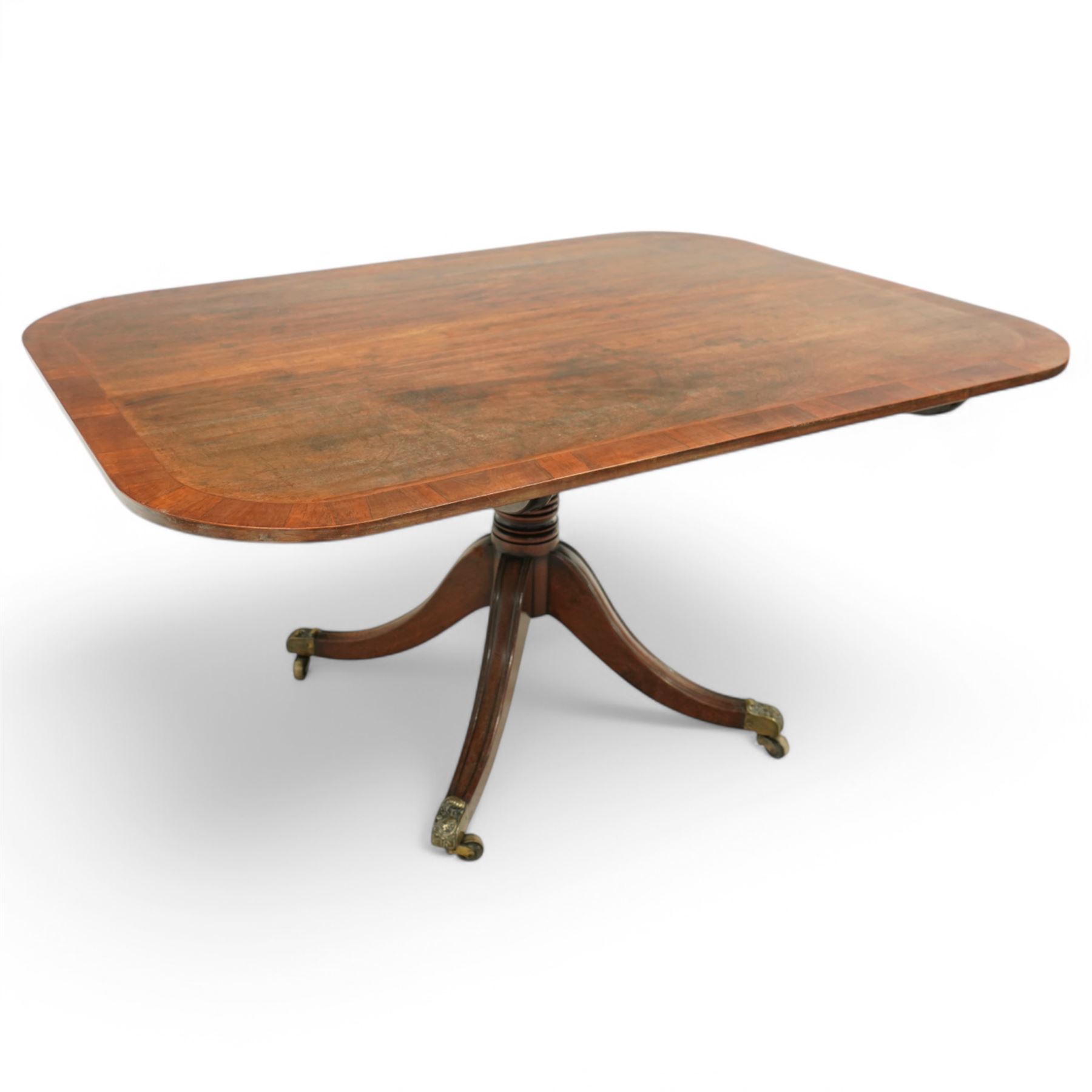 Early 19th century Sheraton style mahogany and rosewood pedestal breakfast table, the rectangular top with rounded corners and crossbanded edge, raised on a turned column support with reeded detail, terminating in four splayed legs with brass caps and castors