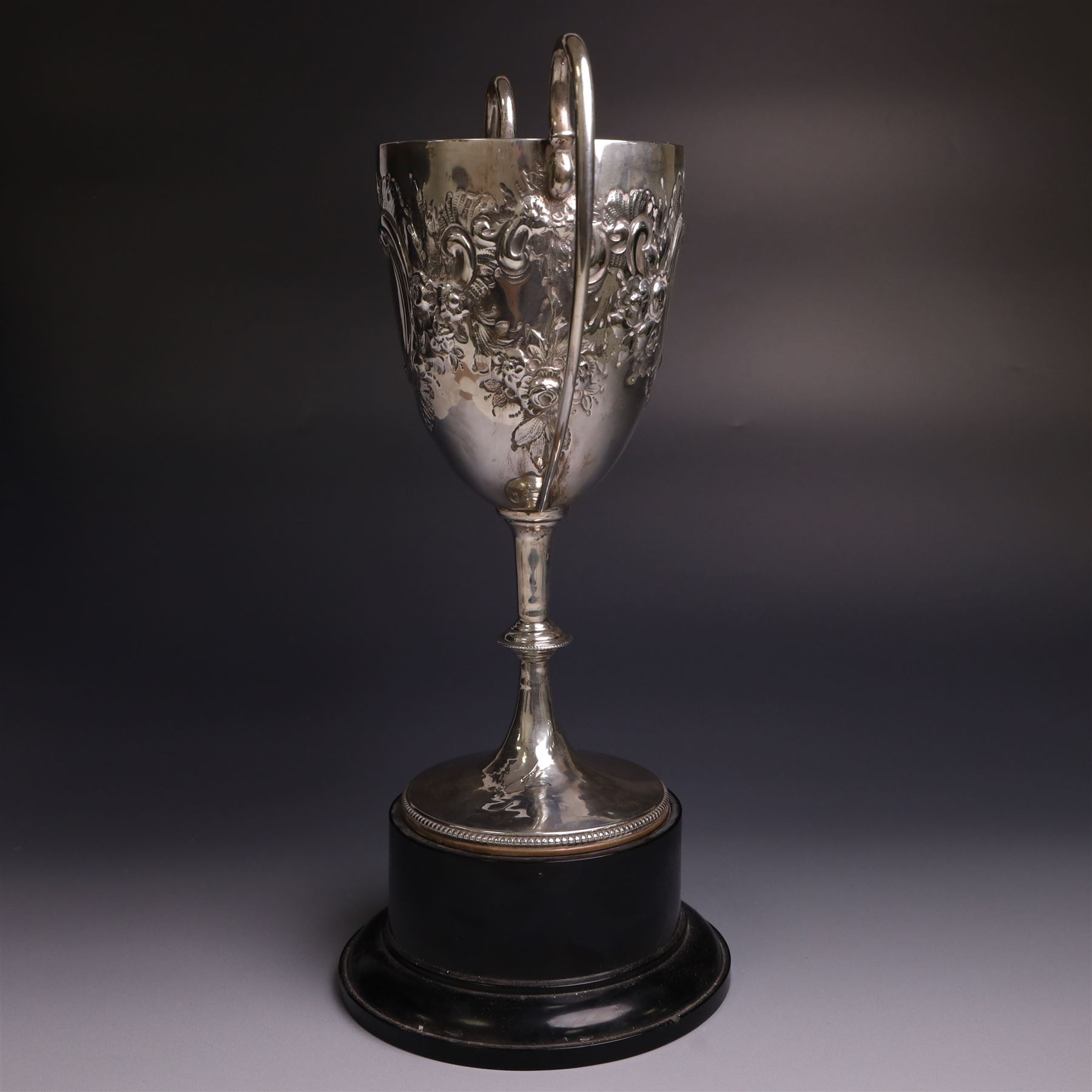 Edwardian silver twin-handled trophy, with embossed floral and scrolling decoration, upon a knopped stem and circular foot, hallmarked Goldsmiths & Silversmiths Co Ltd, London 1903, upon a black plinth, including plinth and handles H32cm