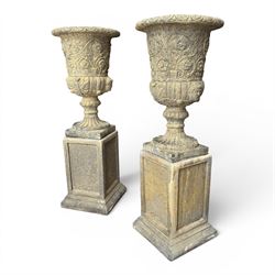 Pair of George III design antiqued composite stone garden urns of campana form, the egg-and-dart everted rim over a waisted body relief moulded with scrolling fruiting foliage and flowerheads, the gadrooned underbelly with twin mask ornaments, the knopped stems on fluted socles, raised on faux panelled square pedestal bases