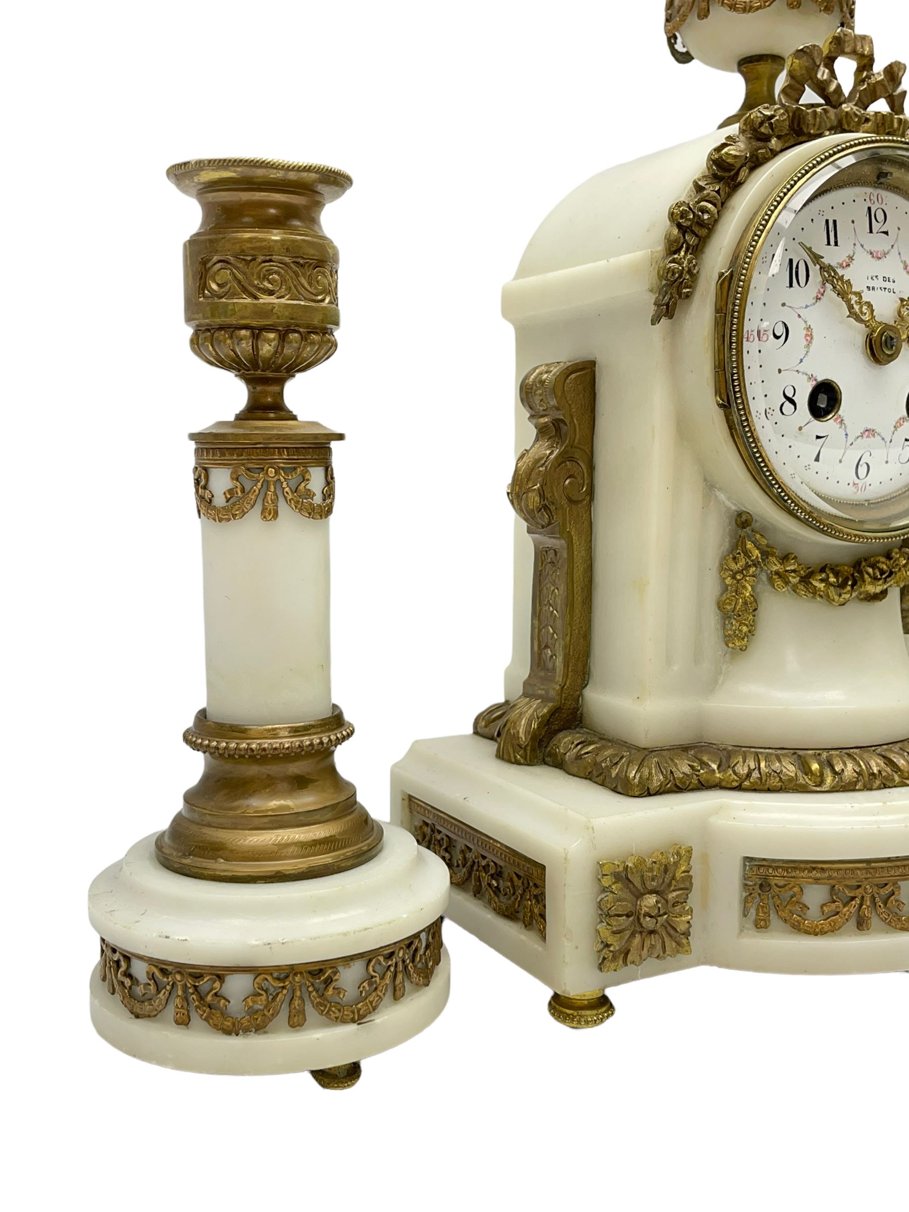 An early 20th century white marble mantle clock in a break front case with an arched top and finial, gilded scroll work to the sides and pierced foliate panels to the front, plinth raised on five bun feet, enamel dial with garland decoration, Arabic numerals, minute markers and Louis XV gilt hands, within a convex glazed bezel, eight-day Parisian striking movement striking the hours and half hours on a bell, with two conforming single light candlesticks. With Pendulum.

