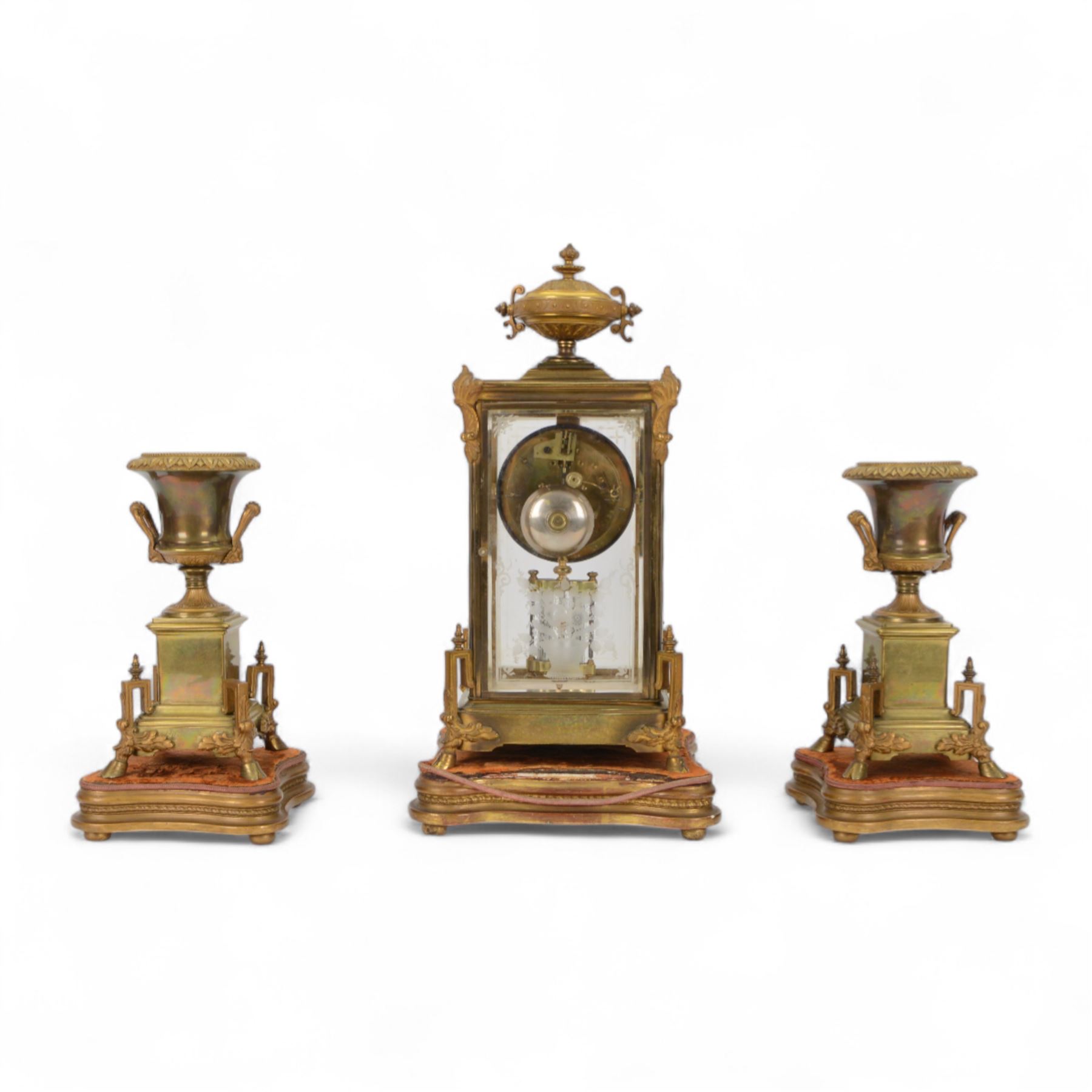 French- Art-deco Edwardian 8-day four-glass clock garniture, with etched glass panels, cast brass decoration to the corners of the case and splayed feet representing cloven hoofs to the base, case surmounted by a large oval urn on a tiered brass platform, two part dial with a recessed brass centre comprising of pierced radial decoration, with steel spade hands and Arabic numerals to the chapter, rack striking movement with a deadbeat Brocot escapement, striking the hours and half-hours on a bell. With a visible twin file mercury pendulum. Garni comprising two large twin handled urns on rectangular pedestals raised on conforming feet representing cloven hoofs, Clock and garni presented on three shaped wooden gesso bases with velvet pads. With key.