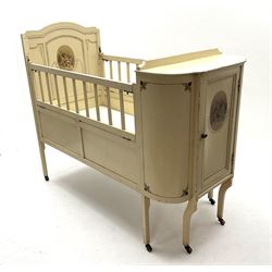 Early 20th century child’s cot, cream painted with applied oval putti scenes, the curved end fitted with cupboard, drop down sides, on square supports with castors