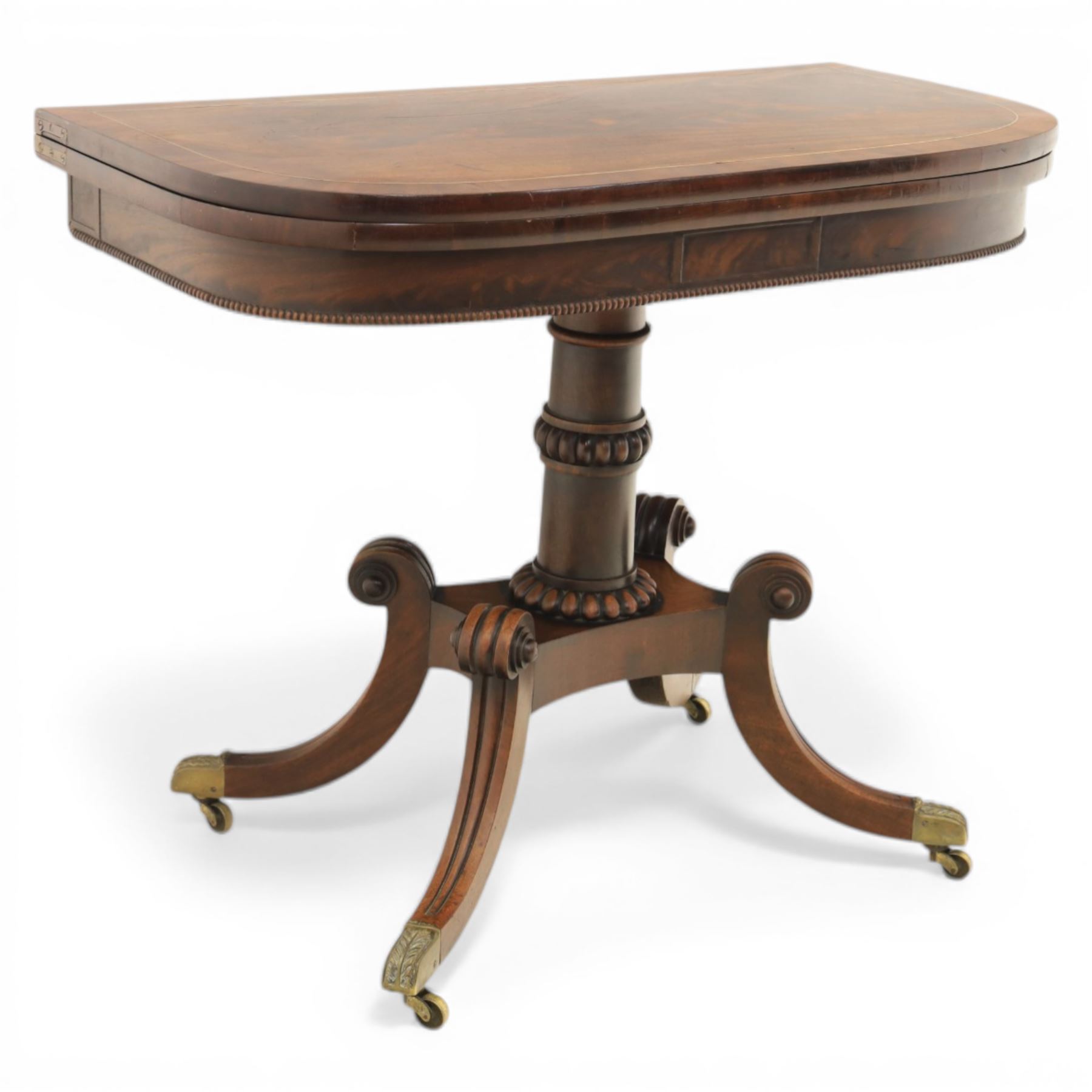 Early 19th century Regency mahogany fold over card table, the rounded rectangular top with brass line inlay enclosing green baize playing surface, crossbanded and strung exterior, with reeded edge above a gadrooned frieze, raised upon a turned column and four reeded sabre supports terminating in brass caps and castors
