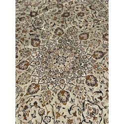 Persian Kashan ivory ground carpet, central medallion decorated with sixteen arranged palmette motifs, within a field decorated with swirling leafy branches and stylised plant motifs, the main border decorated with matching swirling pattern, within floral pattern guard stripes 