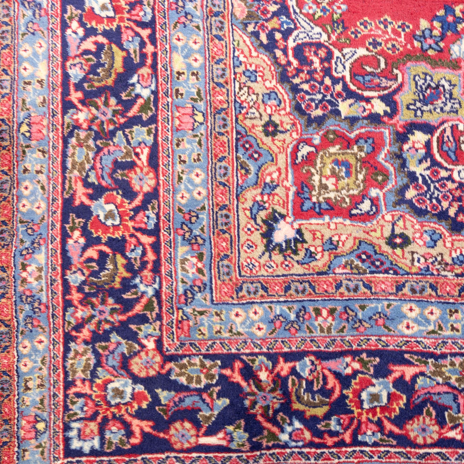 North East Persian Meshed hand-knotted rug, madder red field with central multi-lobbed medallion in shades of indigo, ivory, and sofa pastel tones surrounded by scrolling floral vines, palmettes, stylized blossoms, floral sprays, and cartouches, framed by Indigo spandrels, enclosed within a broad dark blue main border, flanked by multiple narrow guard stripes, with fringe ends 