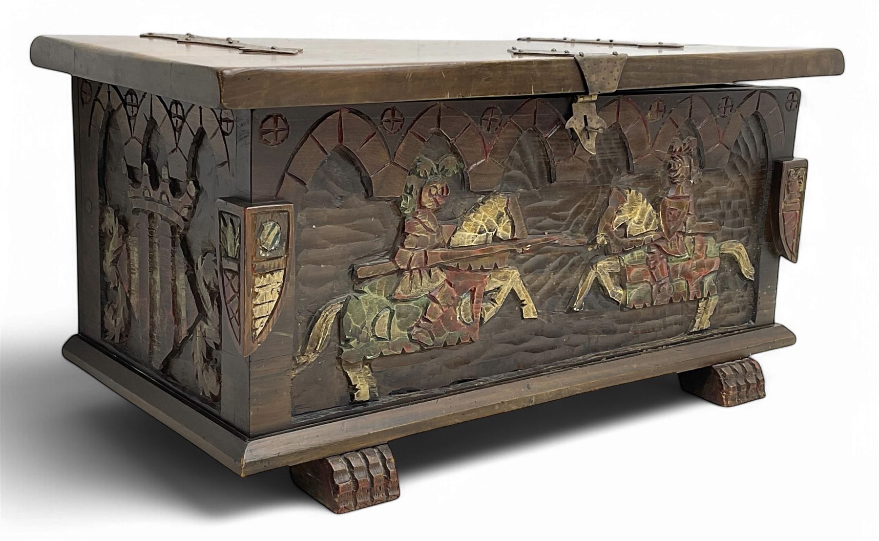 Mid-20th century carved hardwood chest, the rectangular hinged top with decorative strap hinges, the front and sides deeply carved with medieval-style jousting knights, heraldic shields, and architectural elements, raised on shaped block feet