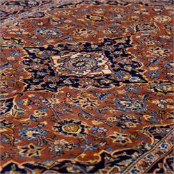 Central Persian Kashan hand-knotted rug,  terracotta field with central shaped ivory and dark blue medallion with palmettes, floral sprays and scrolling vine ornament, with conforming corner spandrels enclosed by a broad navy border of palmettes and flowering vine between multiple guard stripes, with cotton fringe ends 