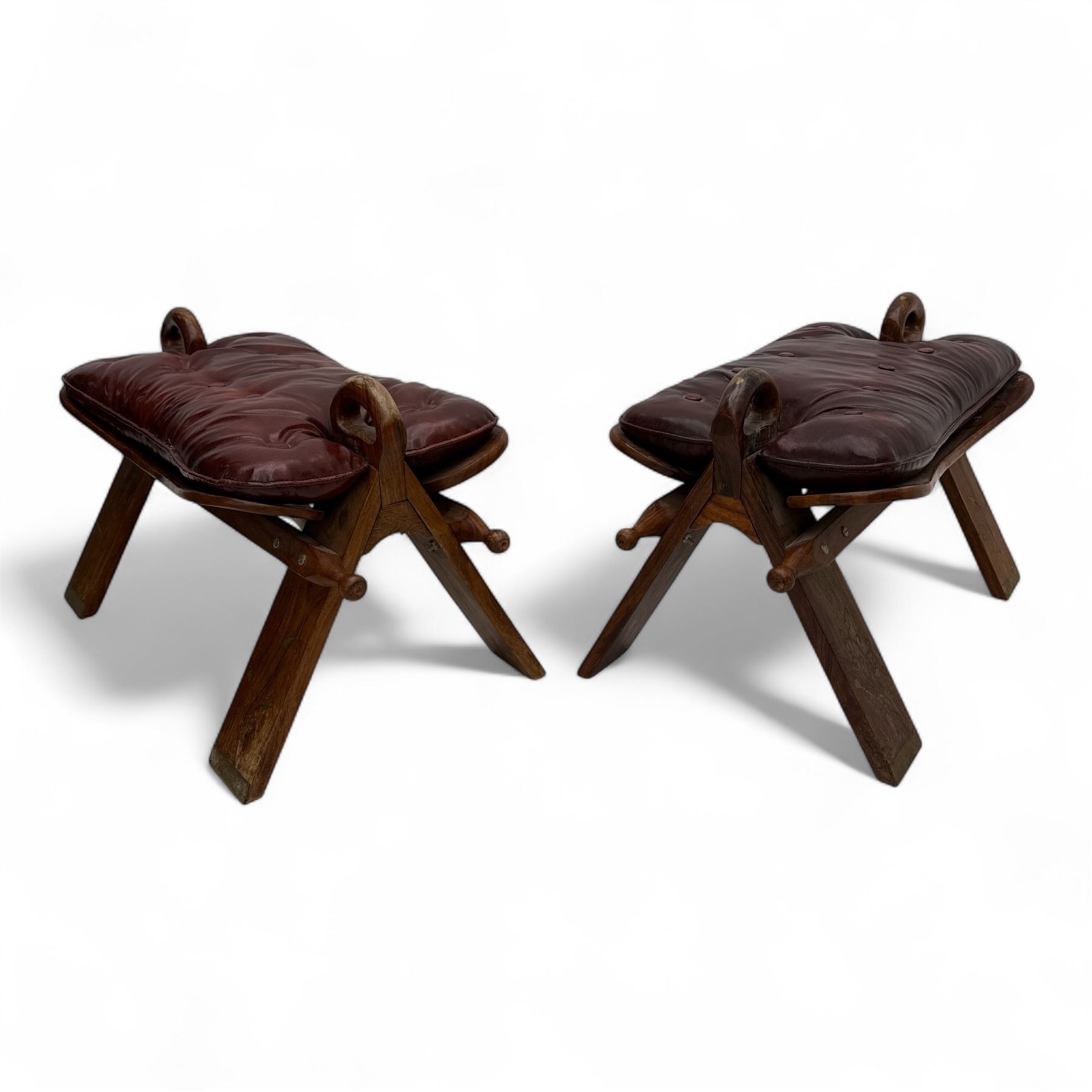 Pair of early 20th century Anglo-Indian camel saddle stools, with deep buttoned red leather upholstered seats and horn-shaped pommels, on X-form supports with turned ball finials