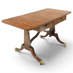 Early 20th century Regency Revival figured mahogany sofa table, rectangular drop-leaf top with rounded corners, fitted with two drawers to each side with brass flowerhead pull handles, raised on twin end supports with splayed reeded legs and brass cups and castors, united by a scrolling and reeded closed stretcher