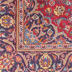 Central Persian Kashan hand-knotted rug, crimson field, profusely decorated with scrolling vines and palmettes, floral sprays around an ivory and indigo lobbed medallion enclosed by spandrels within an indigo border of arabesques between multiple guard stripes  