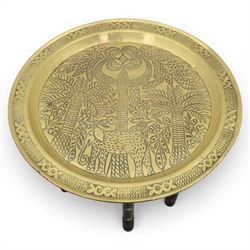 20th century Eastern brass charger topped table, the circular brass trap chased with an elephant, palm tress and flowering vines, supported upon a folding stand with six turned legs with inlaid detail, united by turned supports