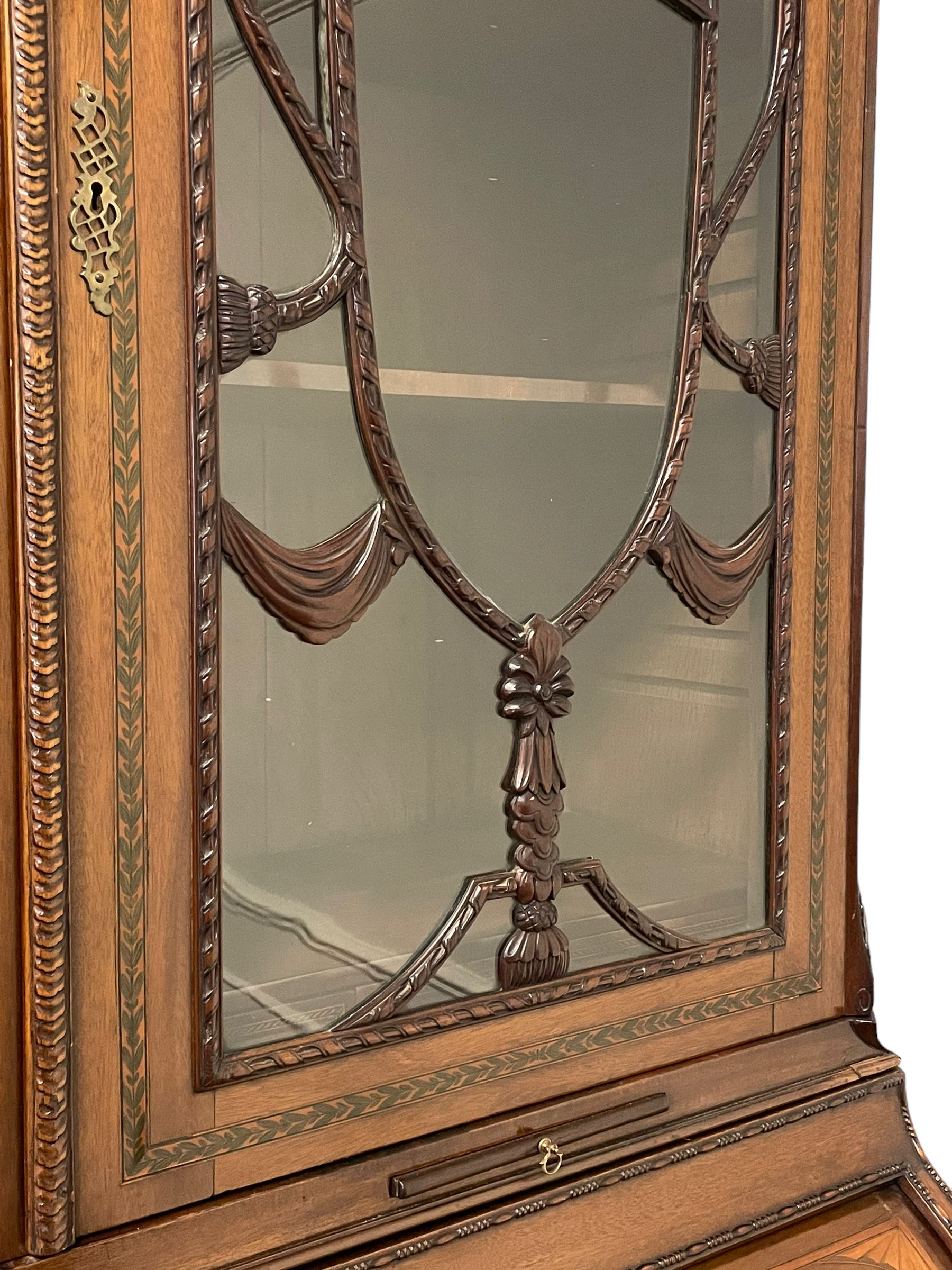 Large and impressive late 19th century Irish mahogany secretaire bookcase, moulded sloped arch dentil pediment decorated with central carved mask surrounded by linen festoons and trailing bellflowers, canted and fluted uprights with carved acanthus leaf upper and lower brackets, enclosed by two astragal glazed doors in the form of trophies with twist ribbon carved mouldings, inlaid with fanned spandrels and trailing leaf painted satinwood band, cylinder roll top inlaid with central fan motif and fanned spandrels, pull-out action revealing stepped fitted interior, central tambour roll cupboard flanked by two Composite pilasters and a combination of drawers and pigeon holes, fitted with three break-front drawers, matched canted and fluted corners with acanthus leaf carvings, on square tapering and fluted spade feet