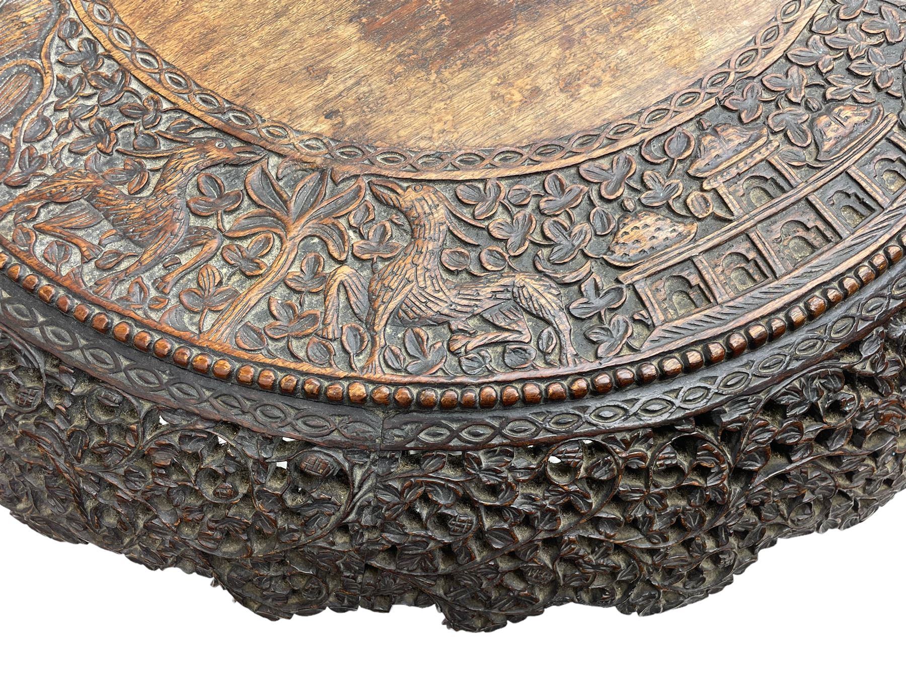 Late 19th century Anglo-Indian carved hardwood occasional table, circular tilt-top carved profusely with mythical animals and foliage decoration, pierced and carved with interlaced leafy branches, leaf carved pedestal supports on a raised platform with further carved and pierced foliage decoration, four carved feet in the form of mythical beasts with gaping jaws 