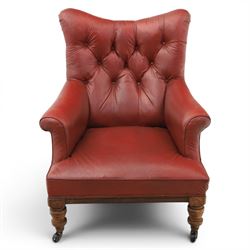 Early 20th century style oxblood red leather Chesterfield armchair, deep buttoned shaped back over padded scroll arms and loose seat cushion, raised on turned mahogany front supports with brass caps and castors