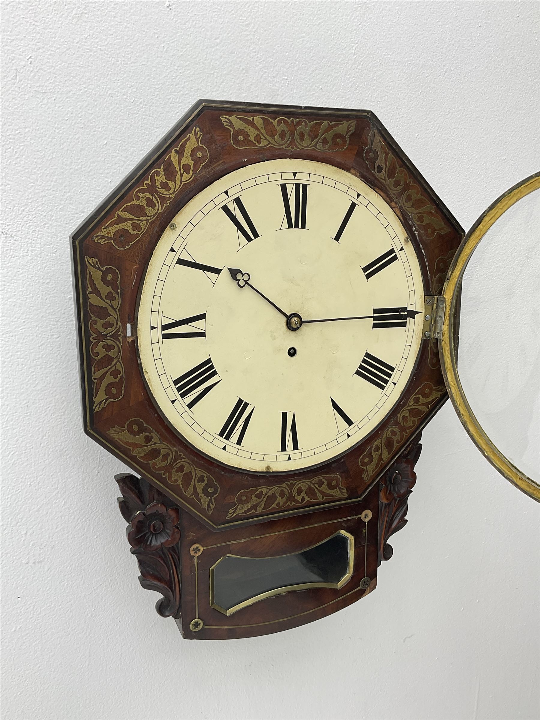 Mid-19th century mahogany veneered eight-day four-pillar single fusee drop dial wall clock with an octagonal dial surround and scroll brass inlay, circular painted steel dial with roman numerals and minute track, matching trefoil steel hands, brass bezel with a flat glass, curved trunk with brass inlaid circles and stringing around the glass pendulum aperture with a brass slip, carved ear pieces in the form of flowers and leaves, with pendulum adjustment door and movement door. With Pendulum & Key