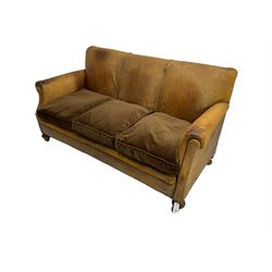 Early-to-mid-20th century three seat sofa, upholstered in worn tan leather with studwork, loose cushions upholstered in brown fabric, on compressed bun feet with castors
