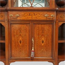 Edwardian Sheraton Revival inlaid mahogany breakfront cabinet, the broken swan neck pediment with central finial inlaid with cartouche and scrolling floral festoons, the central cabinet fitted with astragal-style glazed door with central urn and swag motif, flanked by arched bevelled mirrors over a shelf and gadrooned bracket over paterae inlays, base fitted with marquetry frieze rawer over a panelled cupboard with intricate inlaid scrolling floral designs, flanked by open shelves, raised on square tapering herringbone inlaid supports united by undertier