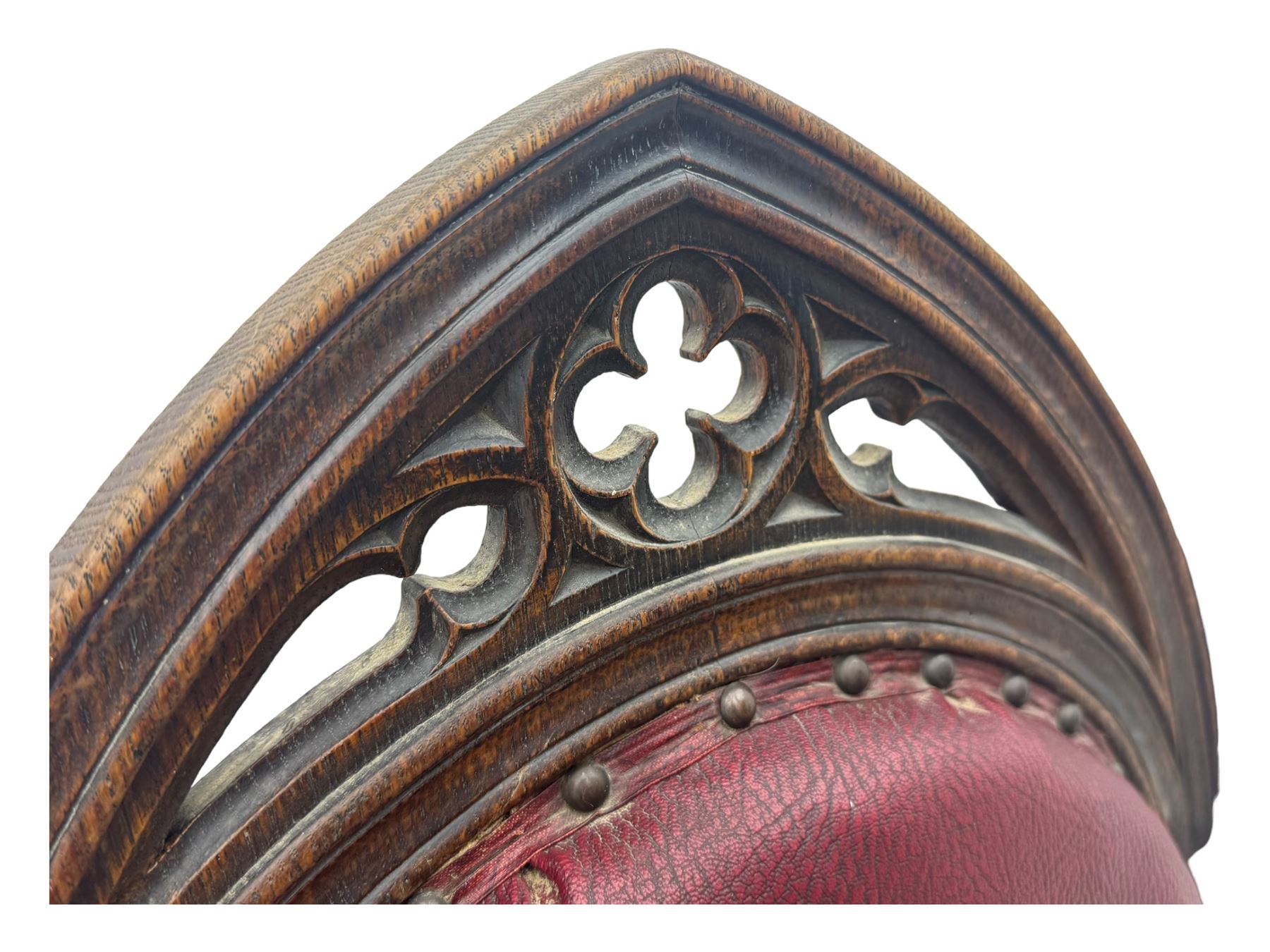 Late Victorian Gothic-revival armchair, arched crest rail carved with tracery and trefoil detail, padded circular back and seat, brass studded burgundy leather upholstery, shaped armrests with scrolled terminals, on square tapering supports