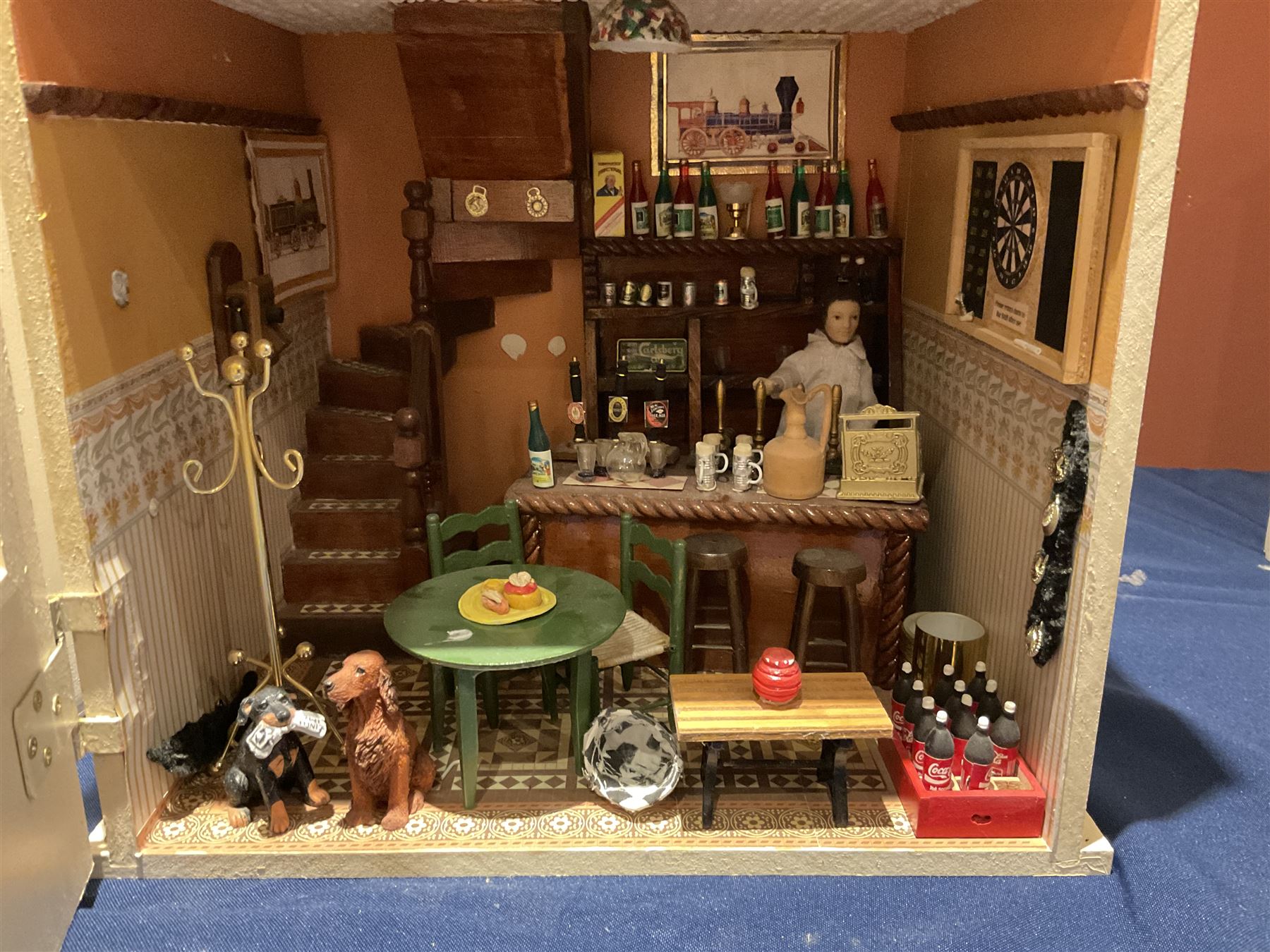 Seagull Pub - scratch-built wooden doll's house as a two-storey gable fronted public house painted in white green and maroon; the hinged front with ground floor veranda and window over opening to reveal two fully furnished rooms with dog-leg staircase and well stocked bar with bar-tender L31cm H62cm D37cm; together with Corner Shop and Dollies Pantry; a similarly painted scratch-built wooden two-storey flat-roofed doll's house as a well stocked ground floor grocery shop with stairs up to two further fully furnished rooms and more stairs giving access to a roof terrace L46cm H57cm D33cm (2)