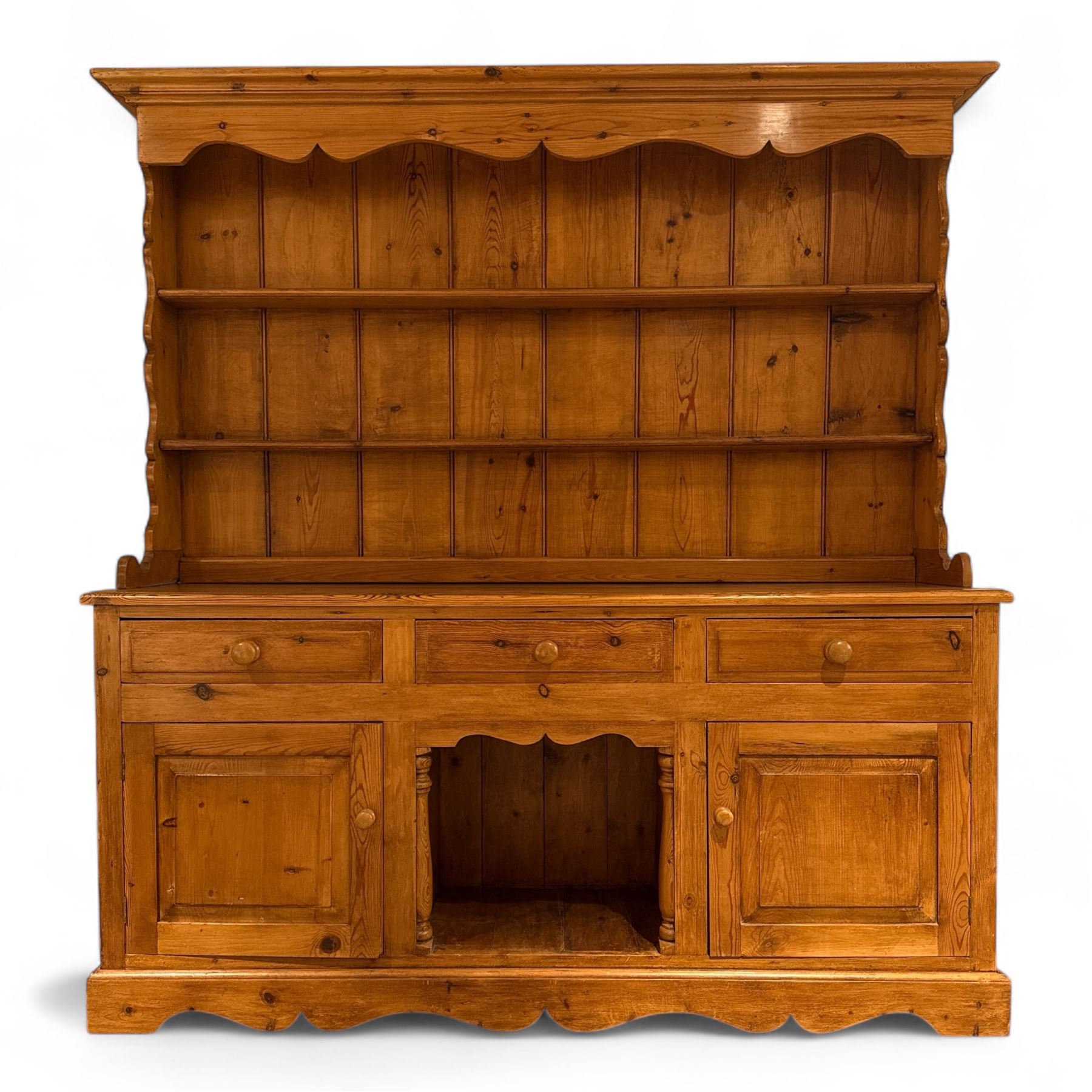 Late 20th century pine dresser, the plate rack with two fixed shelves and shaped apron over panelled back, base fitted with three drawers over two cupboard doors and central open niche flanked by turned uprights, on plinth base with shaped apron
