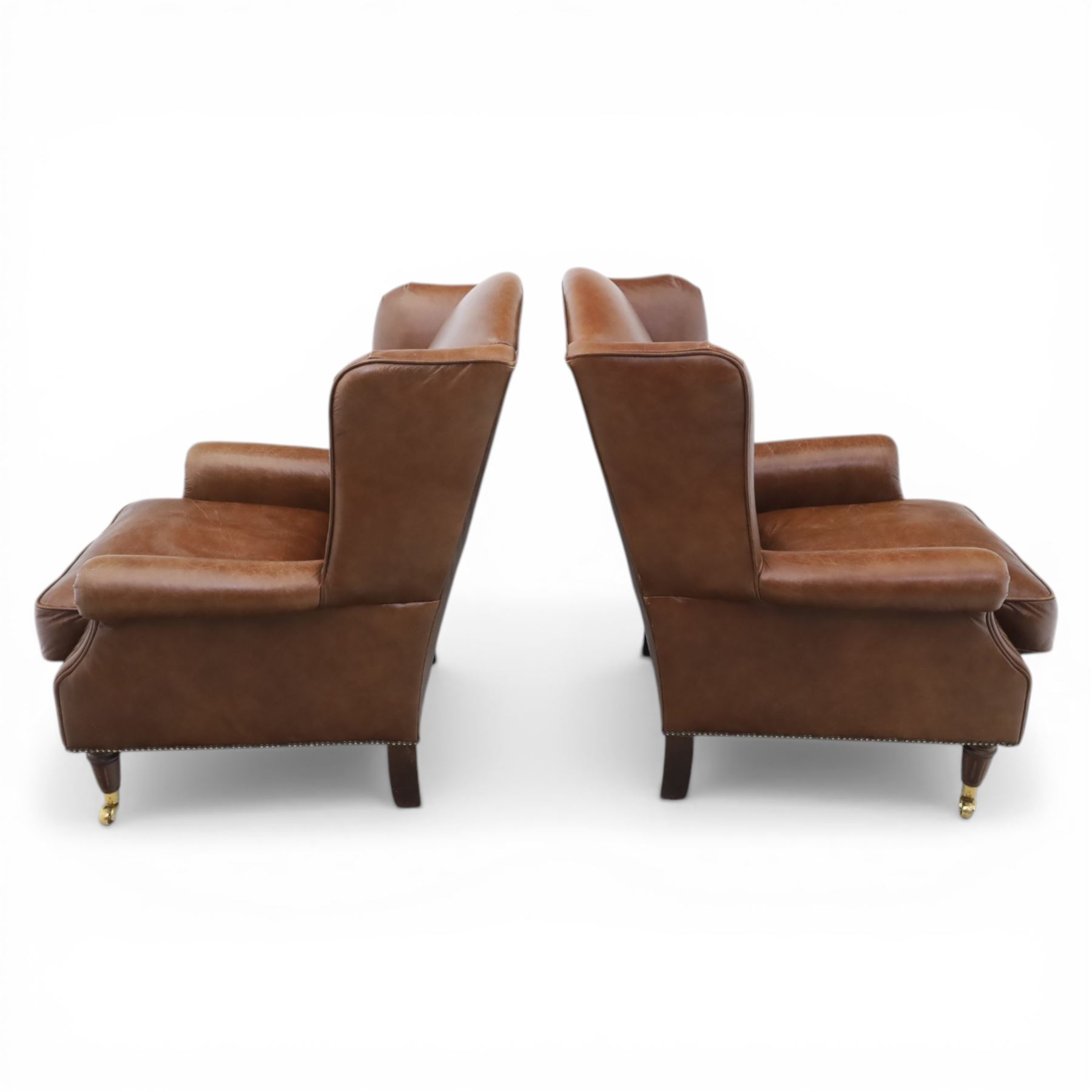 Laura Ashley - Pair of brown leather wingback armchairs, with scrolled arms and loose cushion seat, brass studded trim, on cylindrical tapering legs terminating in castors