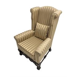 William and Mary style wingback armchair, upholstered in gold striped fabric, on walnut scroll carved supports joined by scroll and foliate carved middle rail, turned H-shaped stretchers, on castors