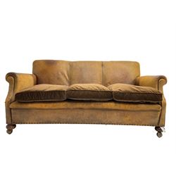 Early-to-mid-20th century three seat sofa, upholstered in worn tan leather with studwork, loose cushions upholstered in brown fabric, on compressed bun feet with castors
