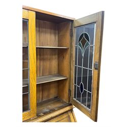 Early 20th century Arts and Crafts oak bureau bookcase, twin leaded and glazed stained glass doors enclosing two shelves, over bureau section with fall-front inlaid with sunburst motif, fitted interior of pigeonholes and shelves, on bracket feet